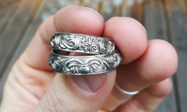 No. 003 Trauringe mit romanischen, Details: Eine Hand hält zwei Trauringe fest und zeigt Details wie einen  überfließender Krug und ein Kreuz in Weißgold.